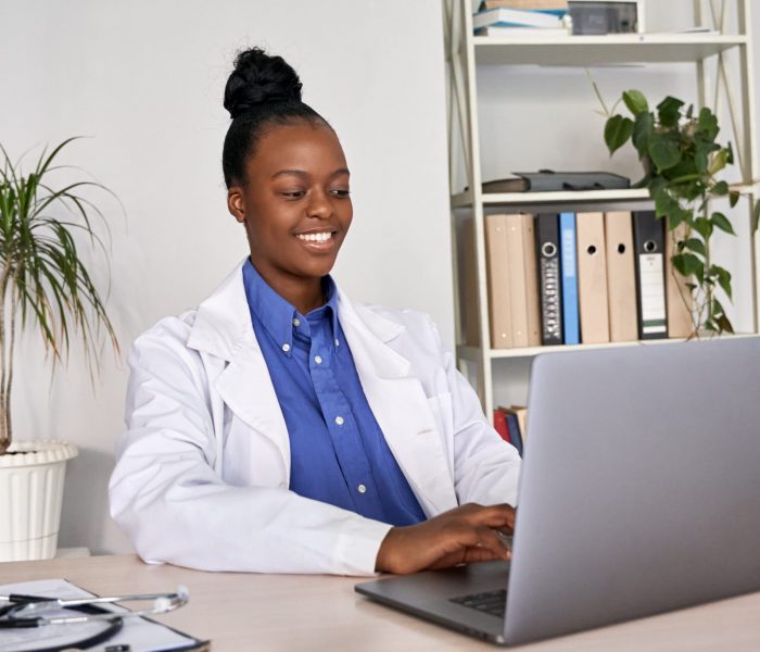 Smiling african american female doctor gp using laptop computer at workplace.