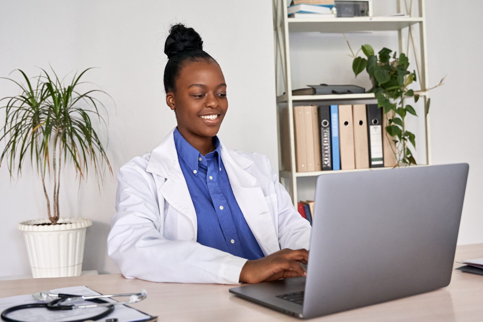 Smiling african american female doctor gp using laptop computer at workplace.